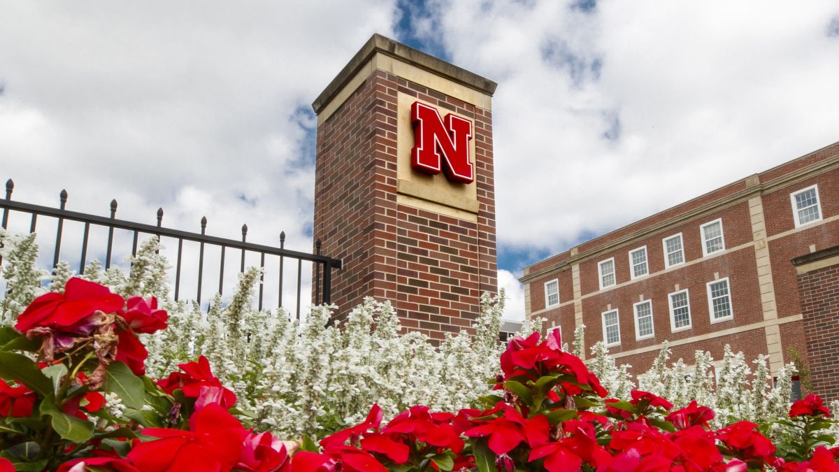 Brick column with a red "N" logo surrounded by flowers, against a cloudy sky and building backdrop.